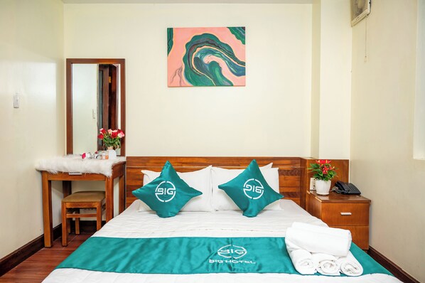 A hotel room with a bed, a wooden nightstand, a mirror, a desk with a chair, and a painting on the wall.