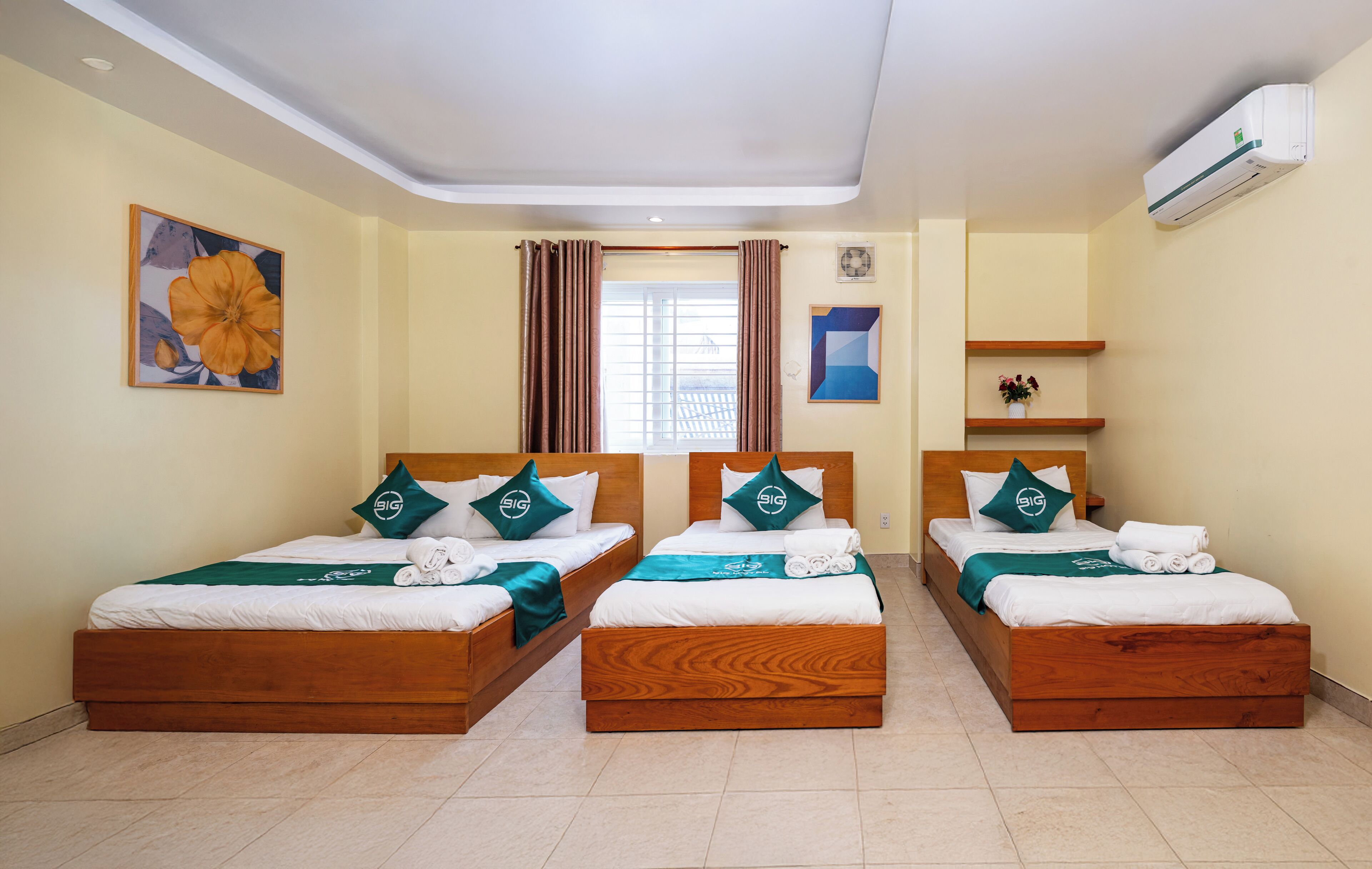 Een hotelkamer met drie houten bedden, elk voorzien van smaragdgroene beddengoed en passende kussens. Er is een raam met gordijnen, een wandairconditioner en een klein plankje met een plantje.