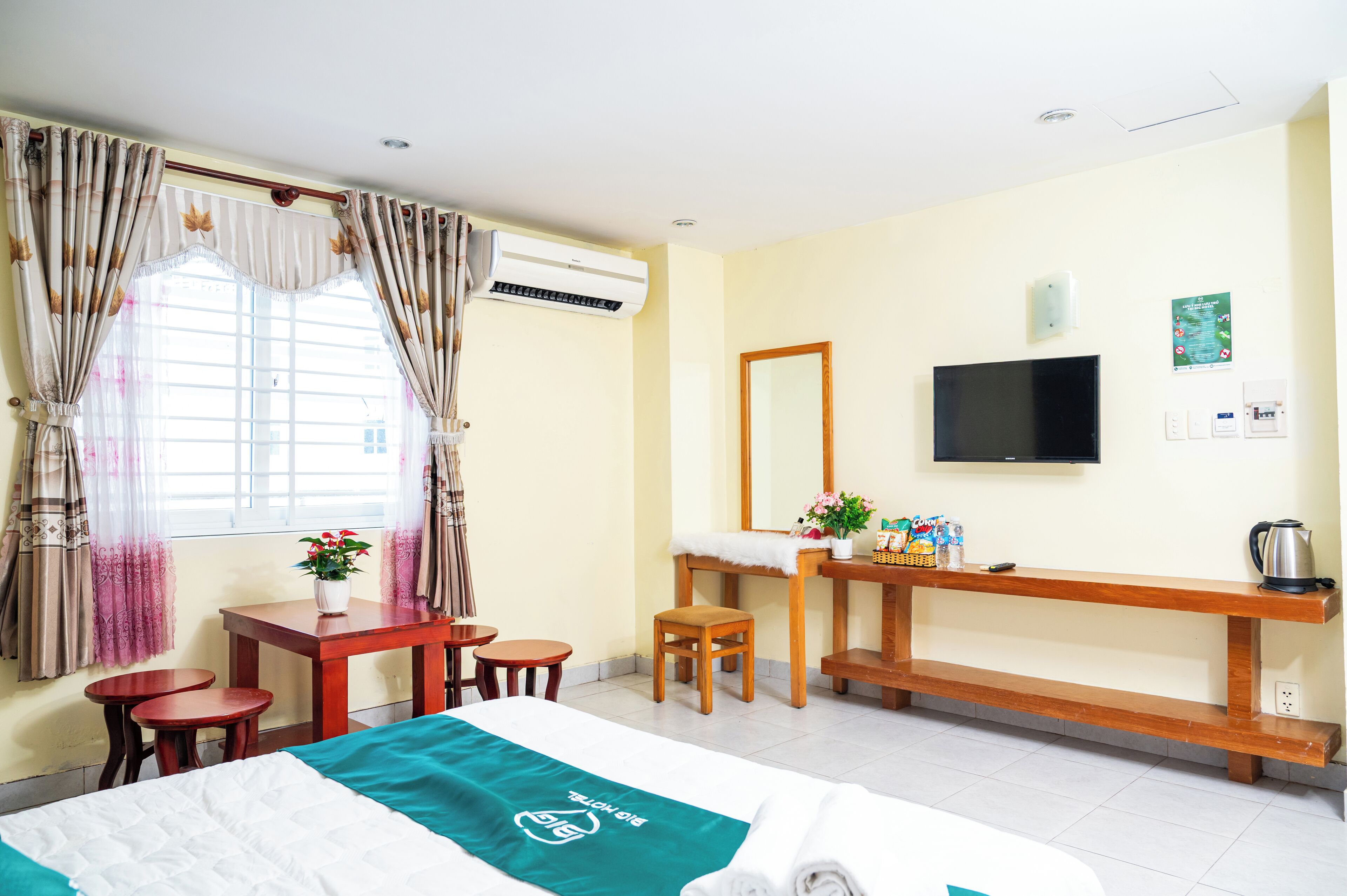 A hotel room with a bed, a desk with a chair, a small table, a mirror, a television, and an air conditioning unit.