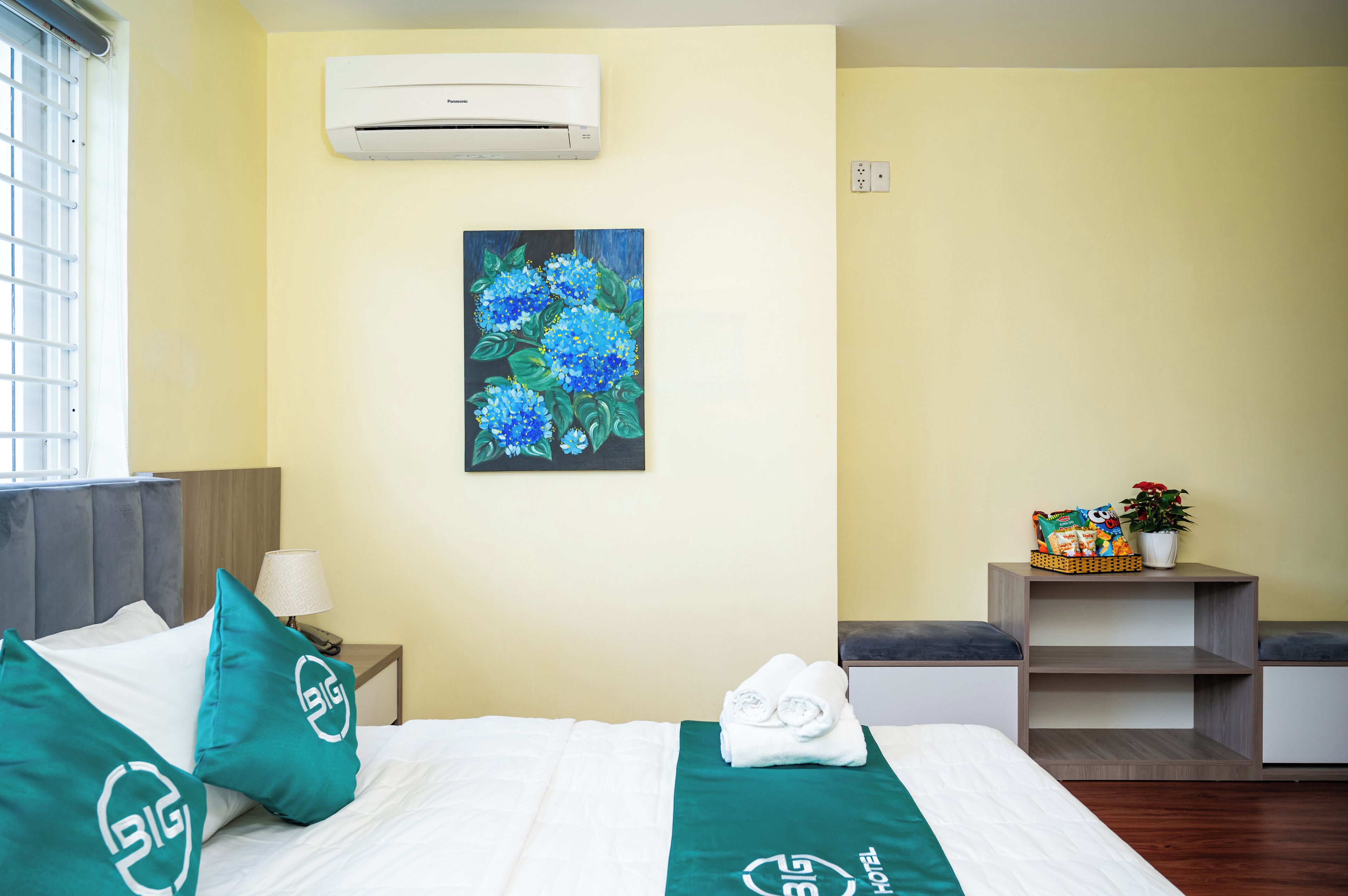 A hotel room with a bed, a nightstand, a wall-mounted air conditioner, a painting, and a small shelf with a basket and a plant.