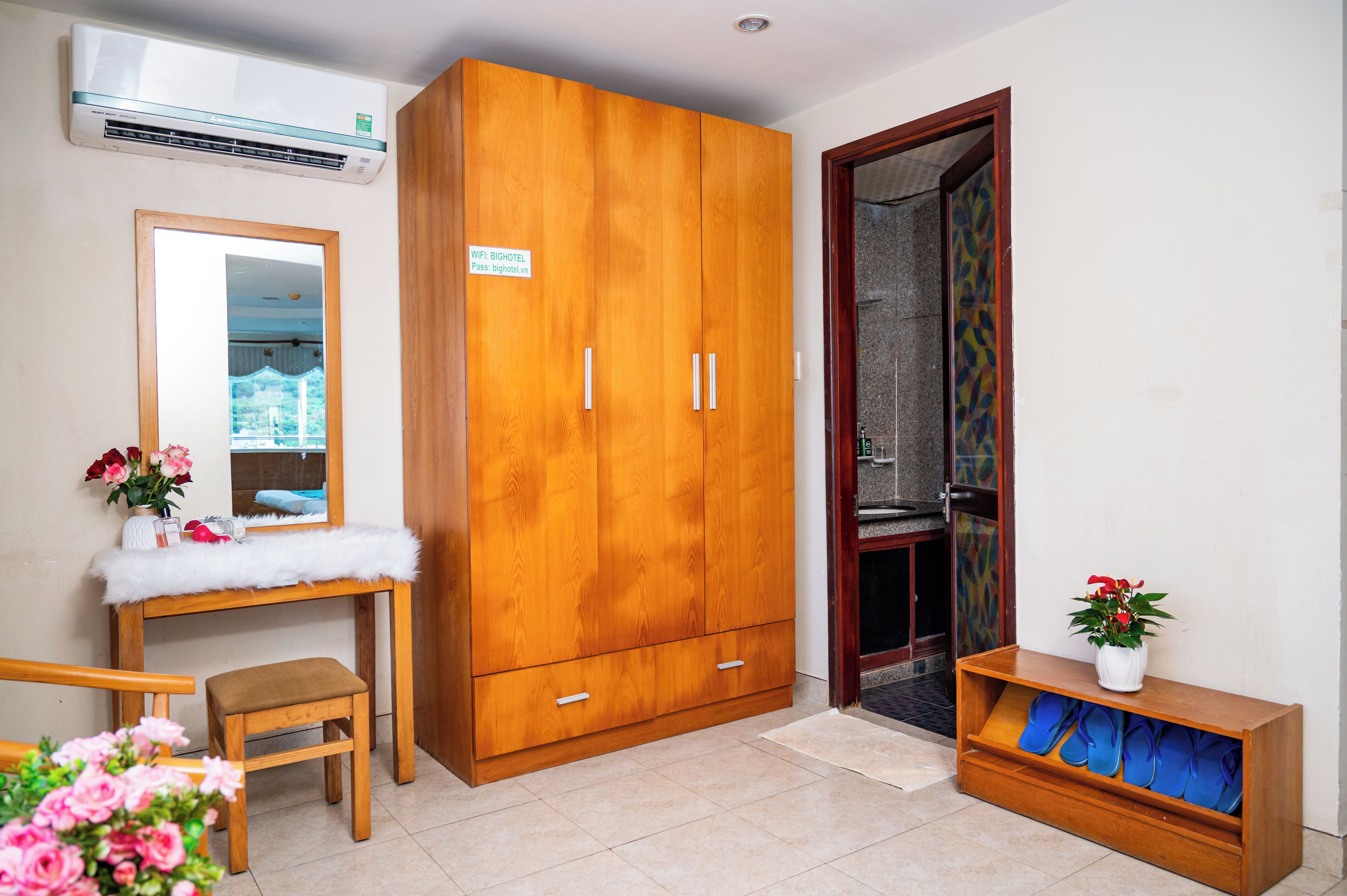 A room with a wooden wardrobe, a mirror, a desk with a white fluffy cover, a chair, and a small shelf with a potted plant.