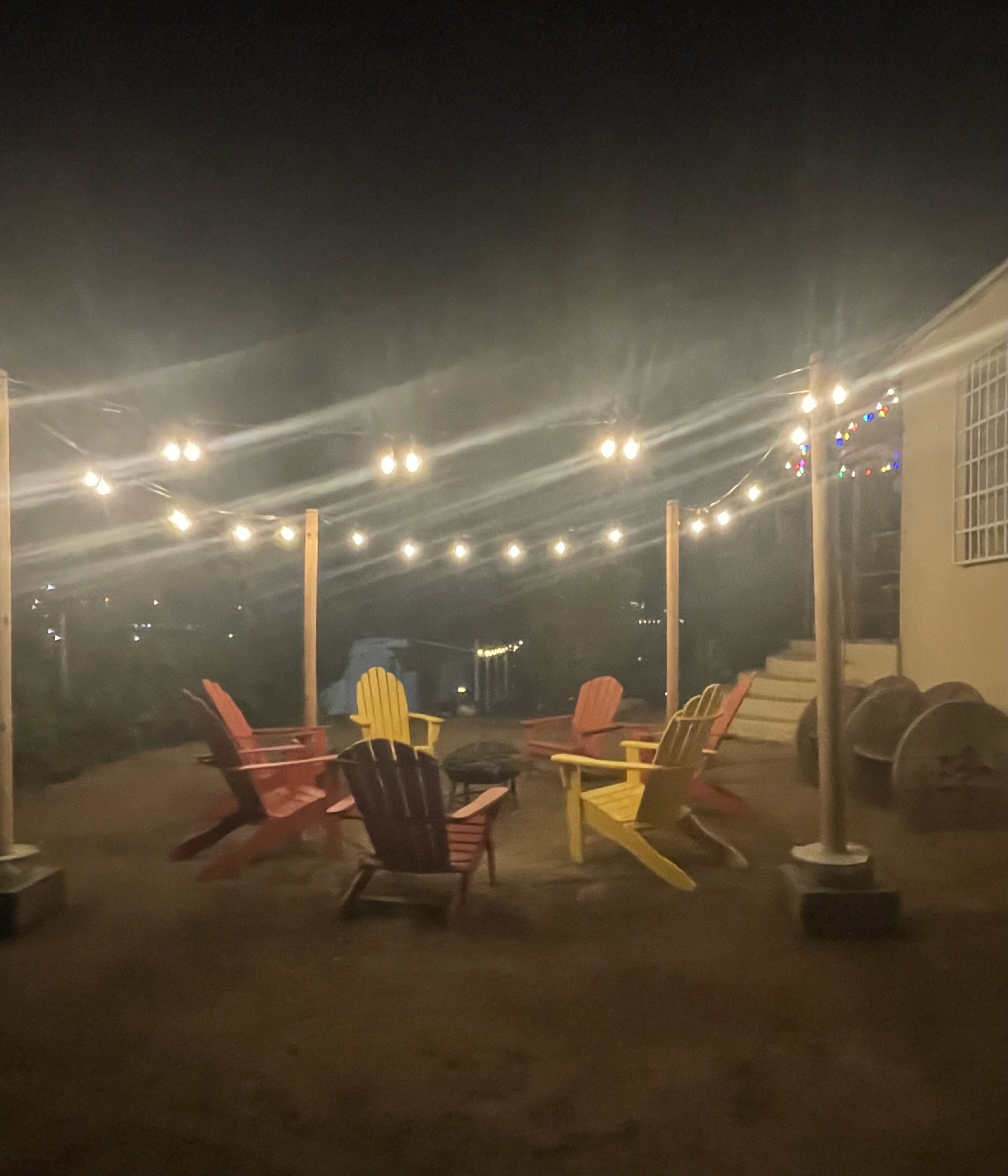 Un área de estar al aire libre con sillas Adirondack y una mesa, luces colgantes y un edificio al fondo.