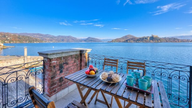 Ein Tisch mit Früchten, eine Schüssel Brot und türkisfarbenen Tassen auf einer Terrasse mit Blick auf einen See und Berge im Hintergrund.