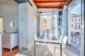Zona pranzo con tavolo in vetro, sedie bianche e vista sugli edifici tramite ampie finestre.