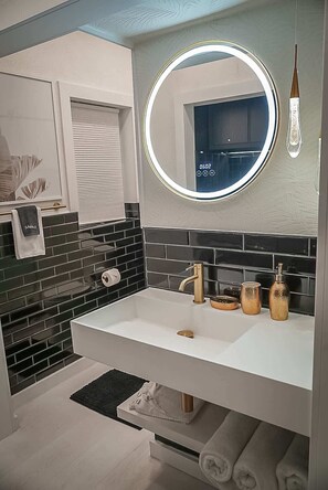 Une salle de bain moderne avec un lavabo blanc, un pare-feu en carreaux noirs, un miroir rond avec un éclairage intégré et un porte-serviettes.