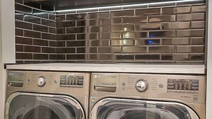 A compact, modern laundry room with two front-loading washers and a stainless steel backsplash above.