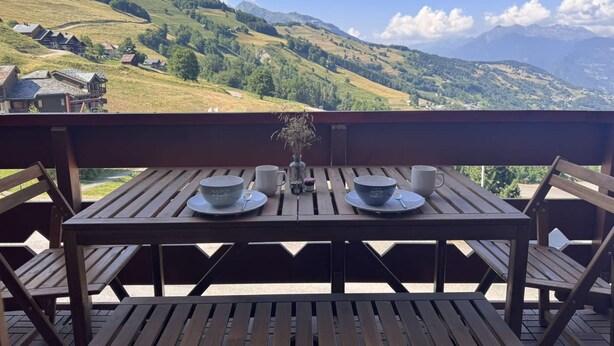 A wooden deck with a table set for two, overlooking a mountainous landscape with houses in the valley. - La Valériane 103 - Beautiful 2-room apartment with an open view of the valley (LES AVANCHERS VALMOREL)