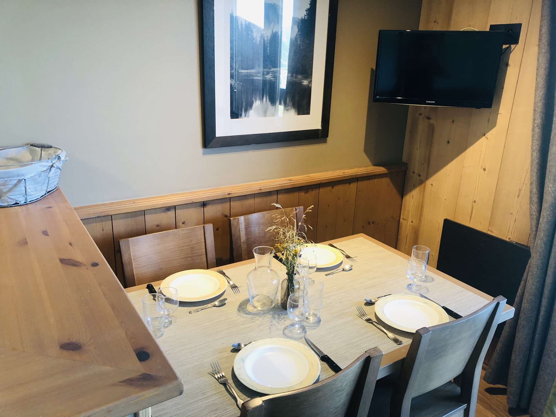 A dining area with a wooden table set for four, a framed picture on the wall, and a flat-screen TV mounted above the table.