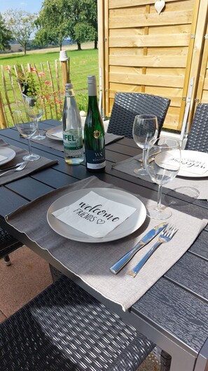 Una mesa de comedor al aire libre dispuesta con platos, vasos, cubiertos y botellas de vino, con vistas a un jardín al fondo.