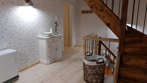 Une pièce avec un plancher en bois, une armoire blanche avec des tiroirs, un radiateur blanc, un panier en osier et un escalier en bois.