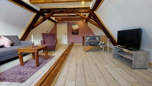 Un salon de style industriel, avec un plafond en pente, des poutres en bois, un canapé, un fauteuil, un meuble pour la télévision et une bibliothèque.