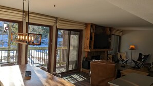 A spacious living area with a wooden dining table, comfortable seating, a fireplace, and large windows offering a view of the outdoors. - Beautiful, Quiet Condo in Keystone! (Keystone)