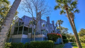 A large, two-story house with a balcony and a covered porch, surrounded by lush greenery and palm trees.
