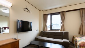 A hotel room with a sofa, a TV mounted on the wall, a wooden dresser, and a window with curtains. - Tabist Business Hotel Osamura (Sabae)