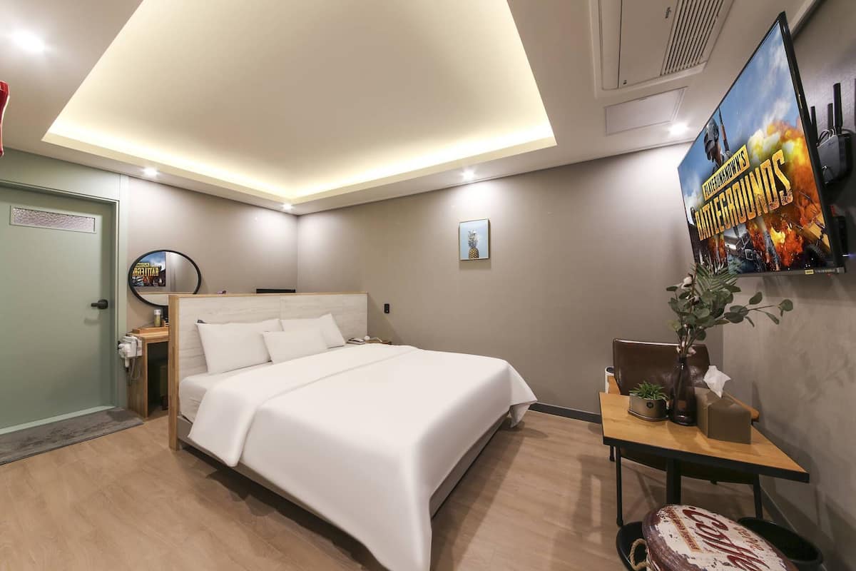A hotel room with a bed, a small table with a plant, a poster on the wall, and a door.