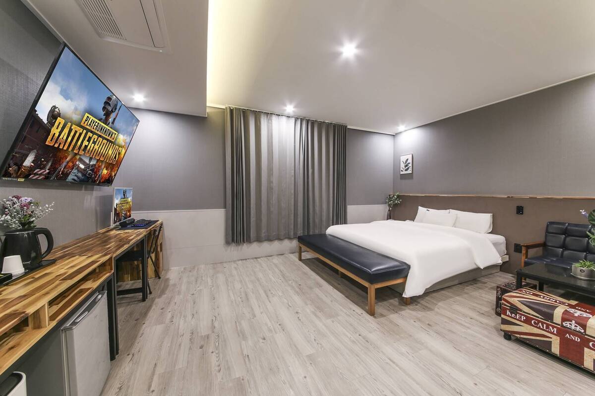 A modern hotel room with a bed, bedside table, desk, and a TV displaying a game.