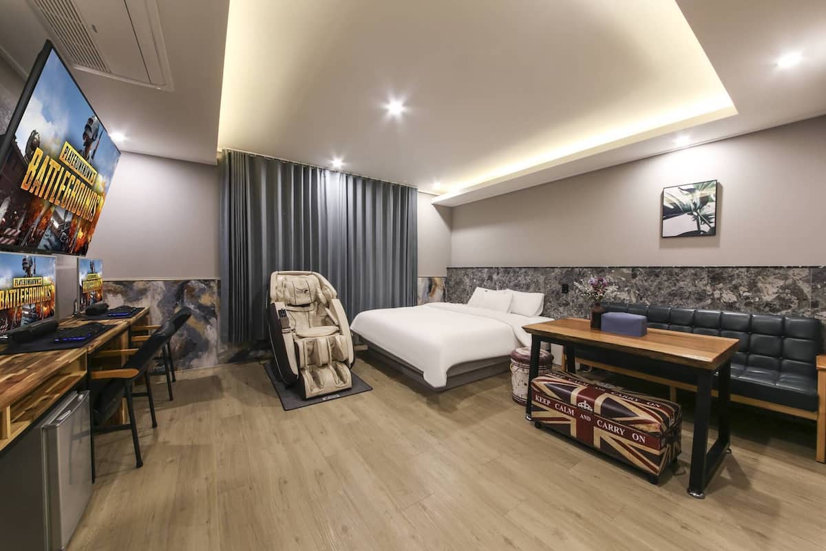A modern hotel room with a bed, a desk with a computer, a gaming chair, a sofa, and a television displaying a game.