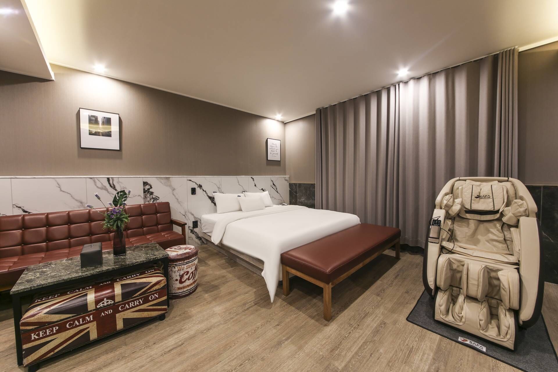 Habitación de hotel con cama, sillón de masaje, sofá de cuero y una pequeña mesa con un florero y un cartel.