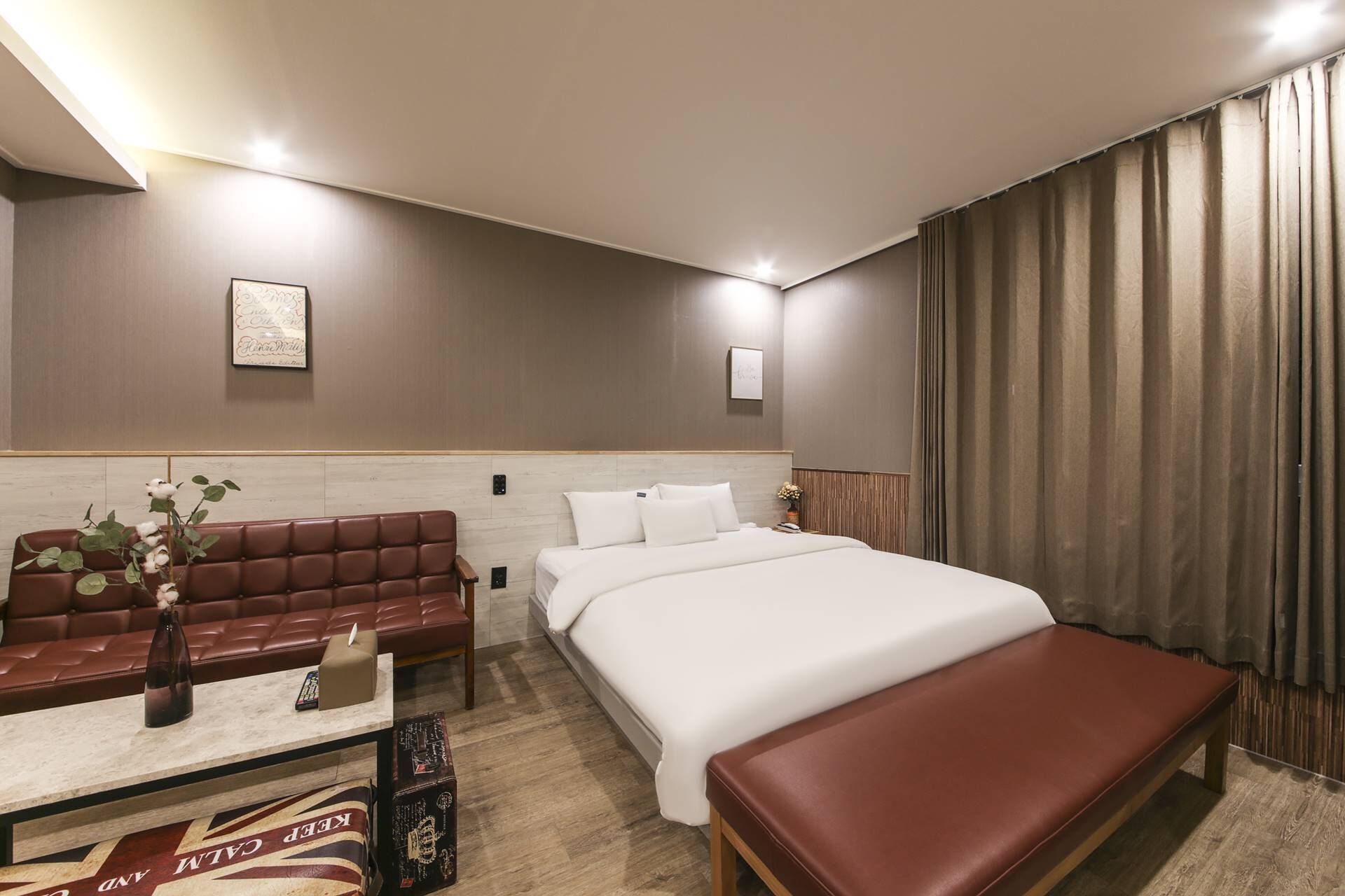 Una habitación de hotel con una cama, un sofá de cuero rojo, una mesita con un jarrón y un banco.