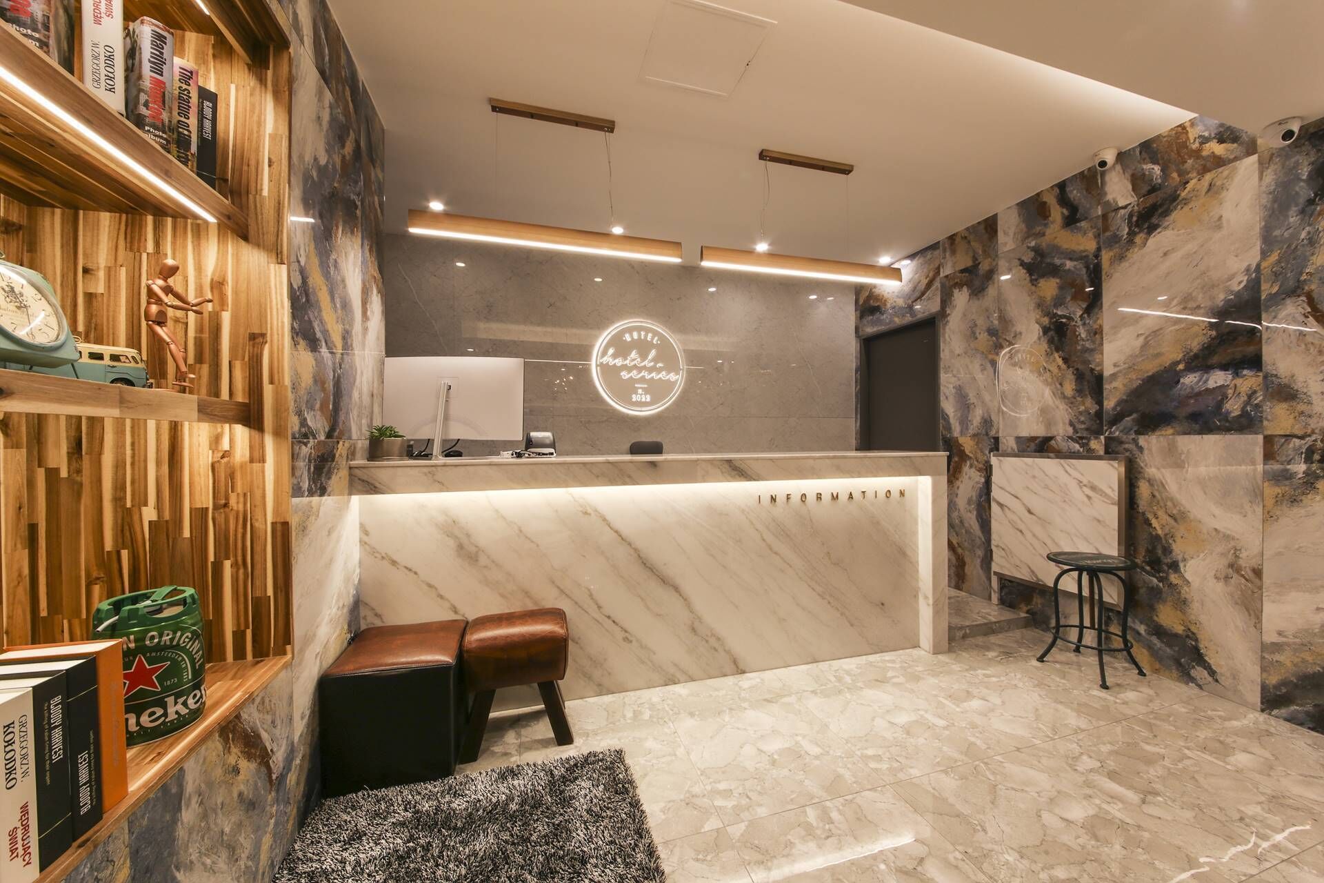 Zona de recepción de hotel con mostrador de mármol, estanterías de madera con libros, un reloj y un letrero de Heineken.