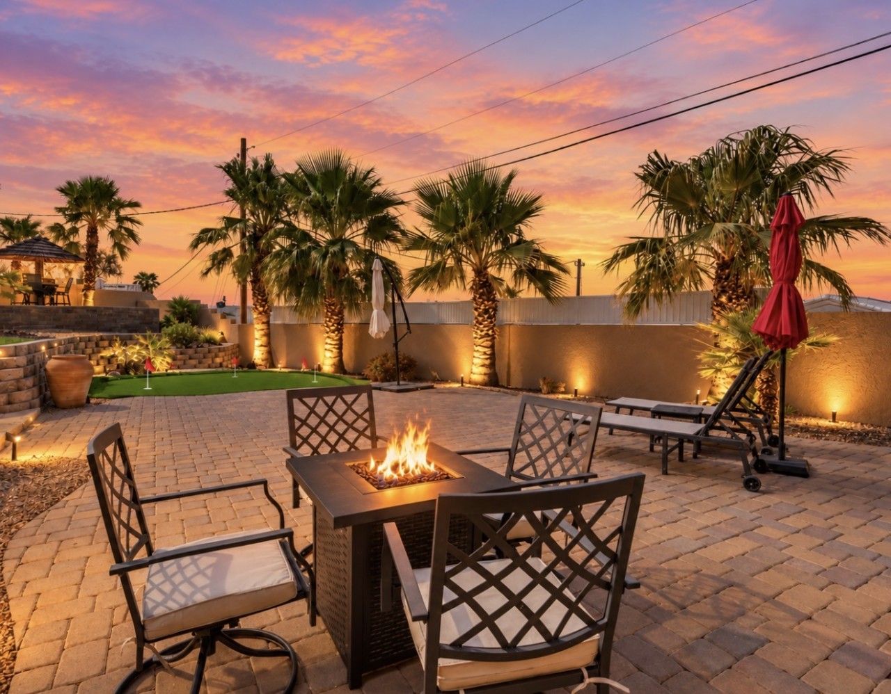An outdoor patio with a fire pit table, lounge chairs, and palm trees.