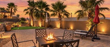 An outdoor patio with a fire pit table, lounge chairs, and palm trees.