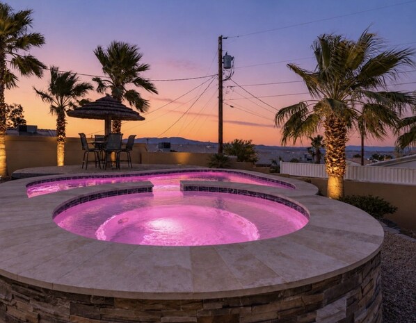 A circular hot tub with a tiled edge, surrounded by stonework, palm trees, and a sunset view.