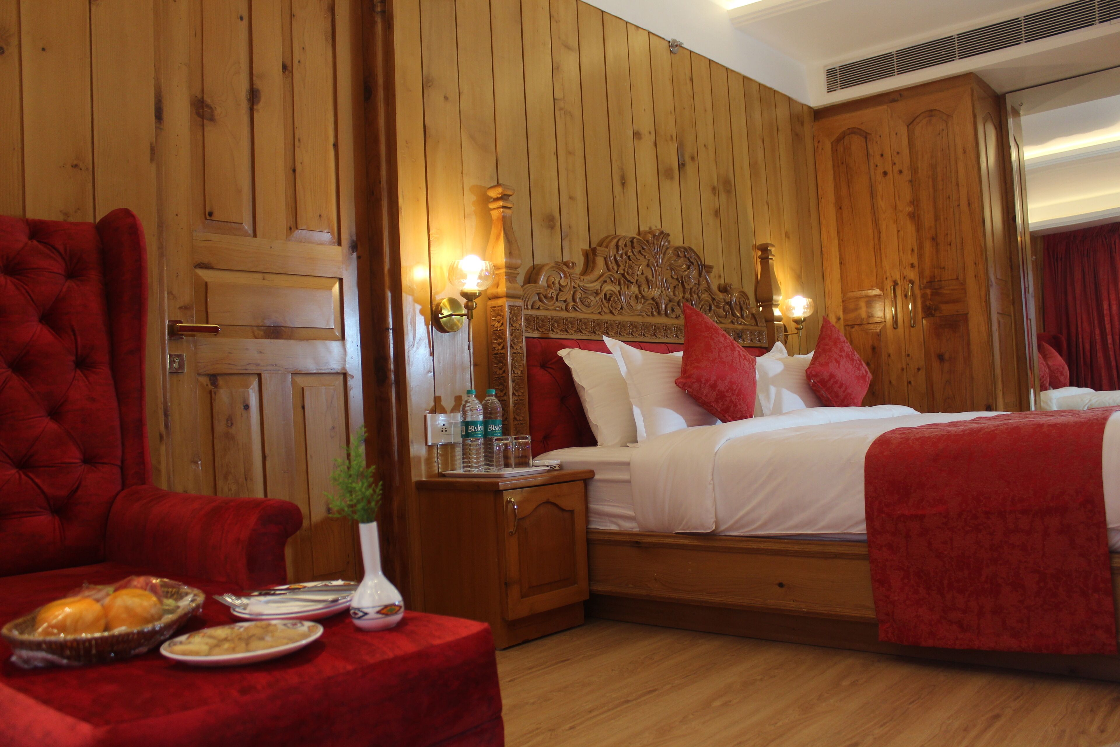 Una habitación de hotel con una pared revestida de madera, un sillón grande tapizado en rojo, una cama con ropa blanca y almohadas rojas, y una mesita con un jarrón y botellas.
