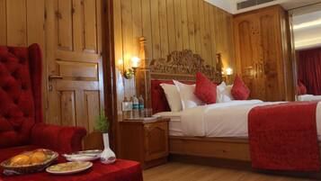 Una habitación de hotel con una pared revestida de madera, un sillón grande tapizado en rojo, una cama con ropa blanca y almohadas rojas, y una mesita con un jarrón y botellas.
