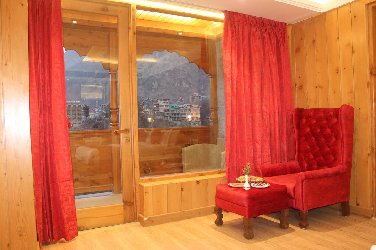A wooden room with a red velvet armchair, a small table with a vase and a plate, and a large window with a mountain view.