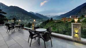 A terrace with tables and chairs, overlooking a mountainous landscape with buildings in the distance.