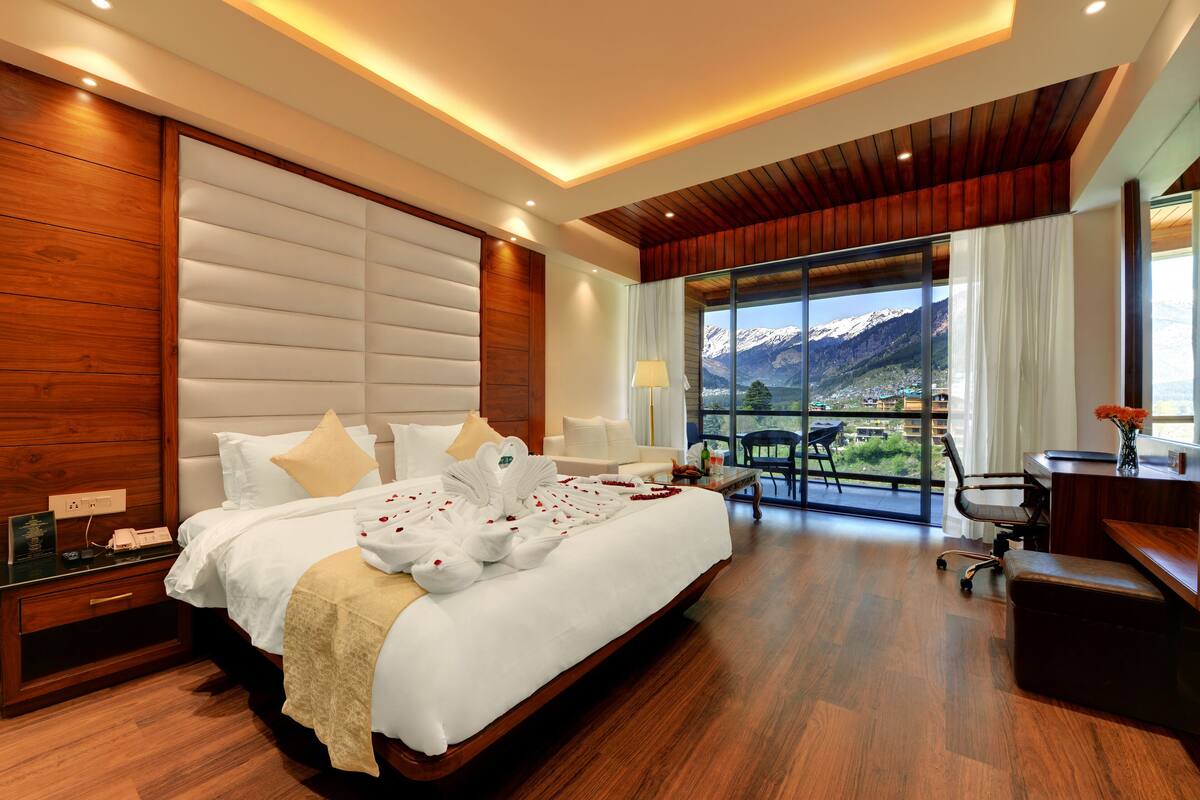 A modern hotel room with a large bed, wooden paneling, a desk, and a view of mountains.