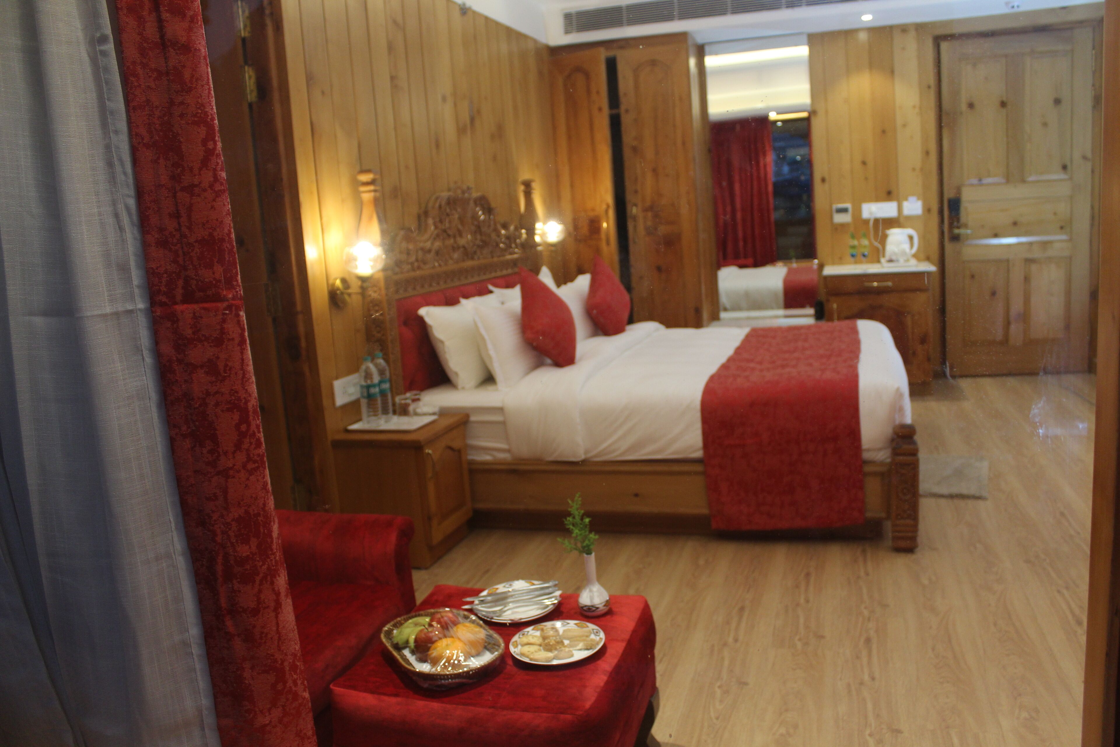 A hotel room with a wooden interior, a bed with red bedding, a small table with plates and a vase, and a nightstand with a lamp.