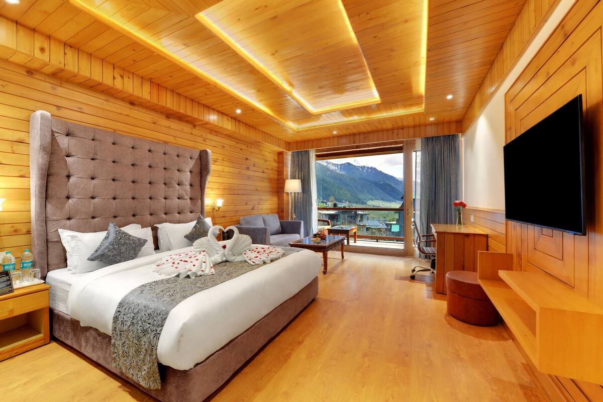 A hotel room with a large bed, a TV, a balcony with a mountain view, wooden walls, and a wooden ceiling.