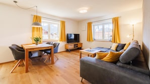 Ein modernes Wohnzimmer mit Holzboden, einem grauen Sofa, einem Esstisch mit Stühlen, einem Fernseher und gelben Vorh ängen.