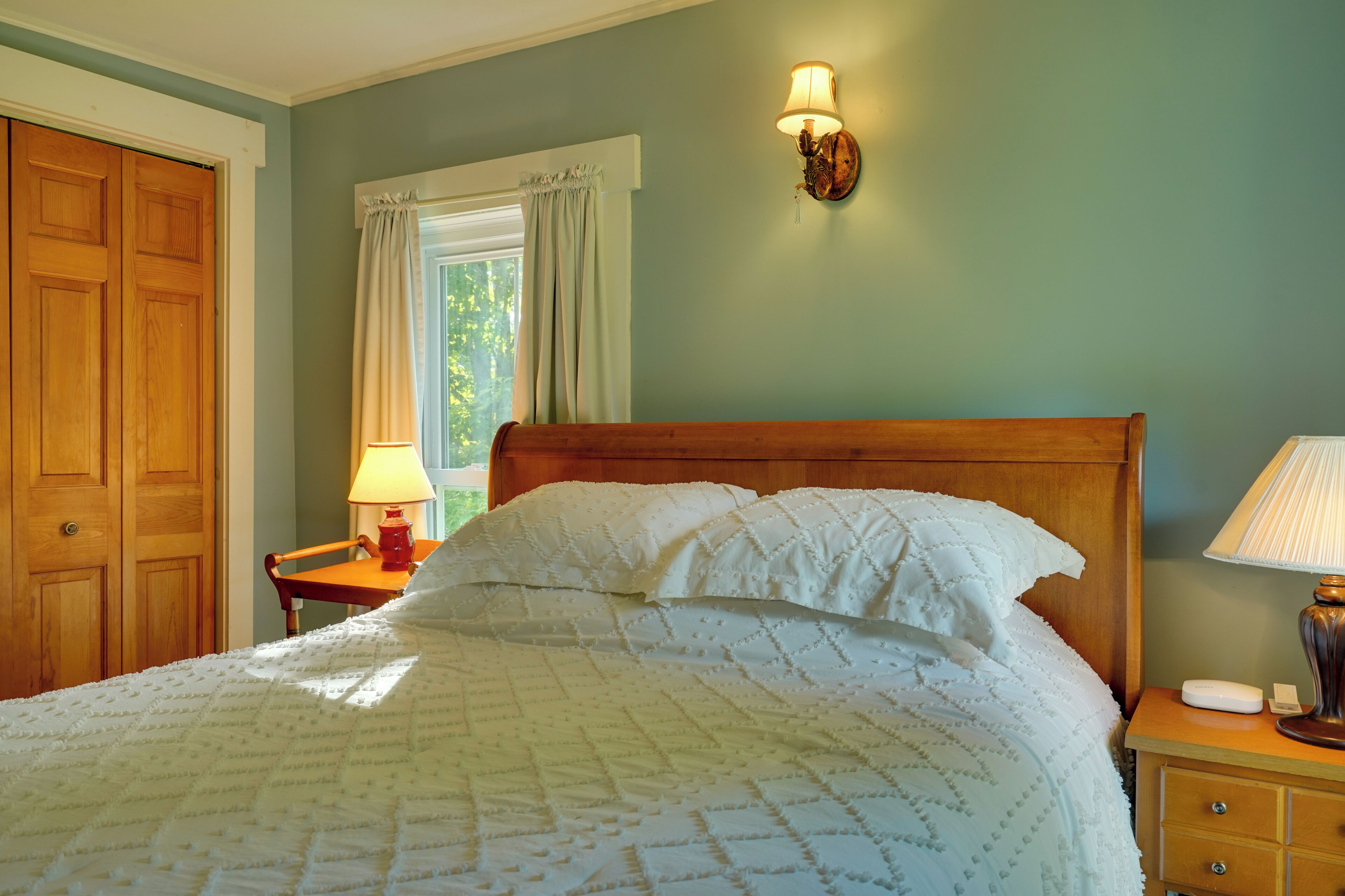A bedroom with a wooden bed, a nightstand, a wall-mounted lamp, and a window with curtains.