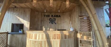 A reception area with a wooden reception desk, a sign reading "THE KAMA", and a bamboo structure in the background.