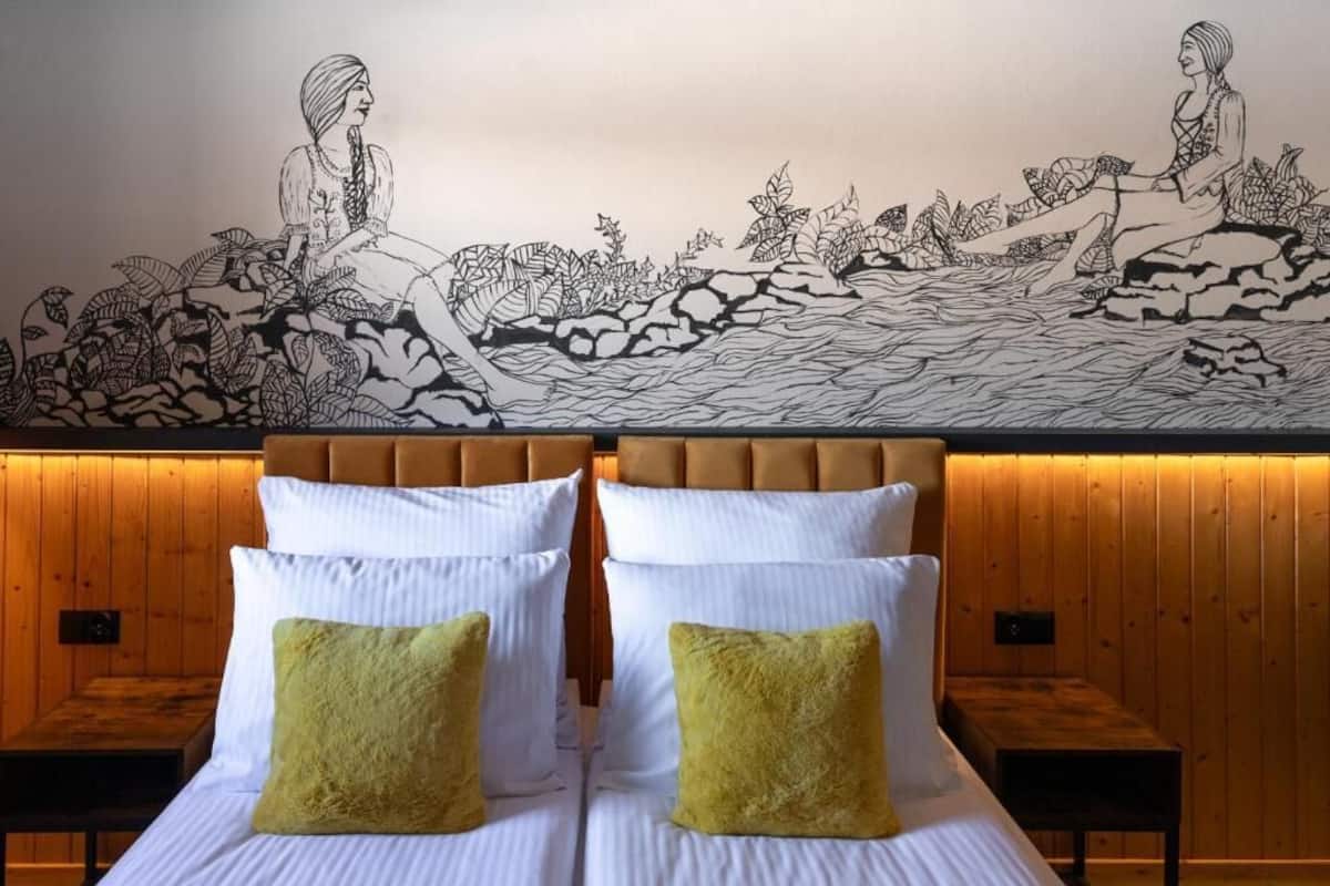 Ein Doppelbett mit weißen Leinentüchern und zwei grünen Kissen, ein hölzernes Kopfteil und ein Wandbild, das zwei Frauen auf Steinen zeigt.