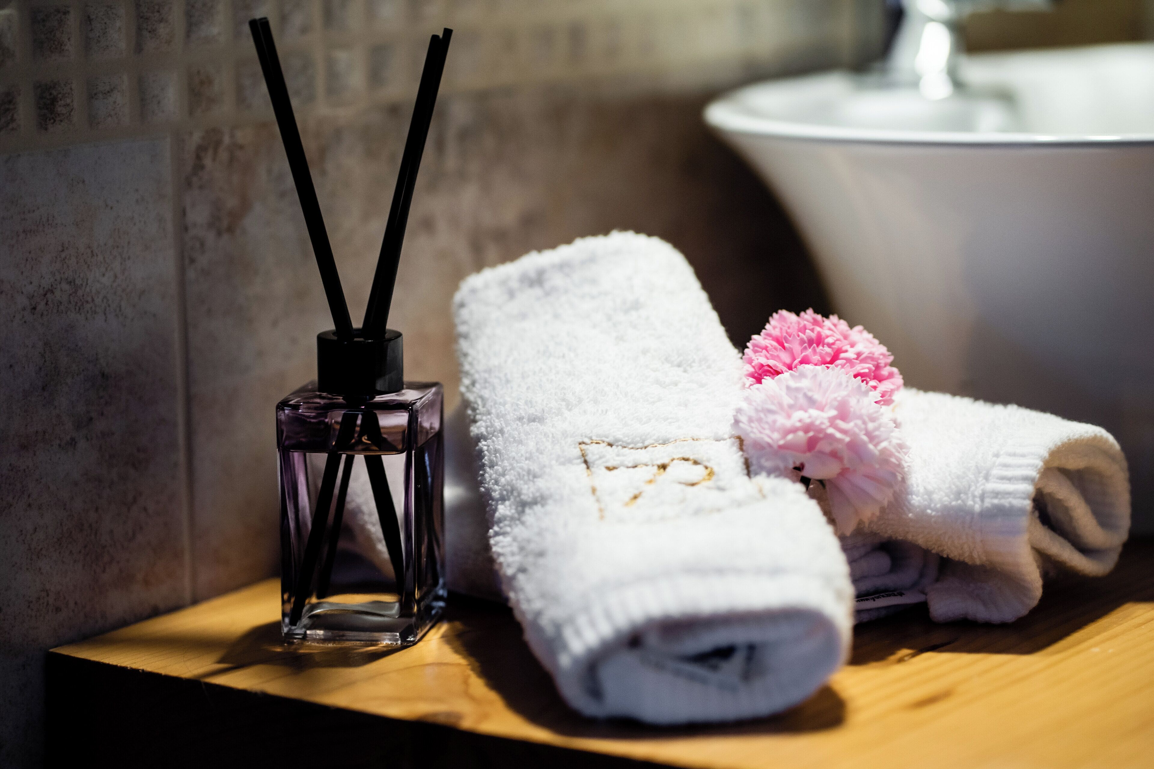 A bathroom with a wooden counter, a white towel with a pink flower, and a glass bottle with black sticks.