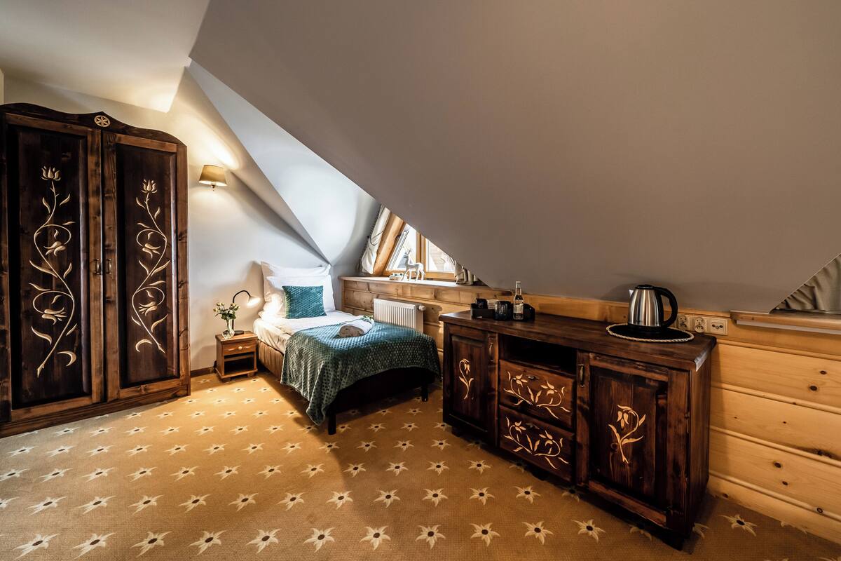 Ein gemütliches Schlafzimmer im Dachgeschoss mit schrägen Wänden, einem Bett mit grüner Bettdecke, einem Holfischschrank mit floralen Schnitzereien, einem kleinen Nachttisch mit Lampe und einem Heizkörper.