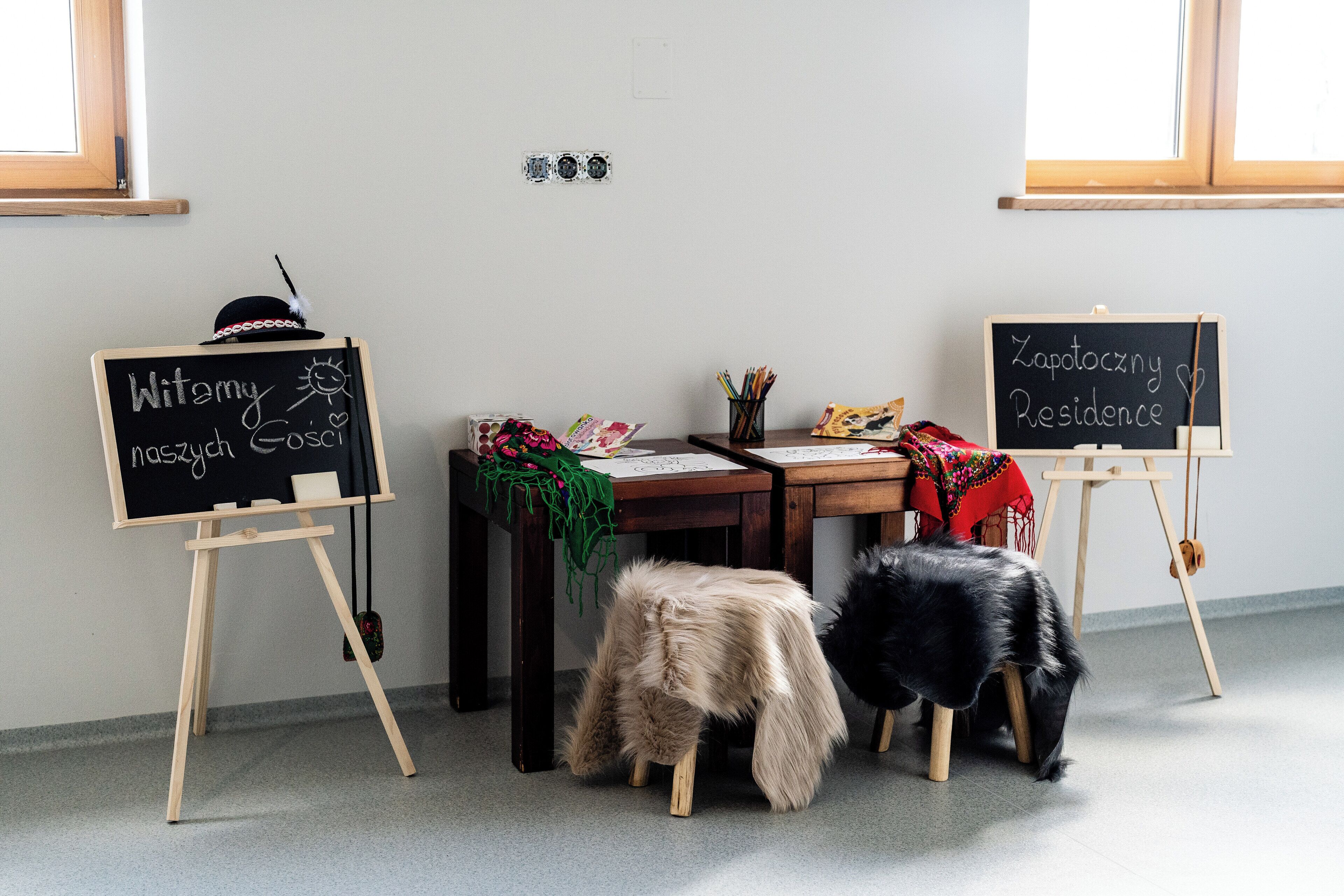 Une pièce comprenant deux tableaux noirs sur lesquels du texte est écrit, un bureau avec du matériel de dessin et un petit tabouret.
