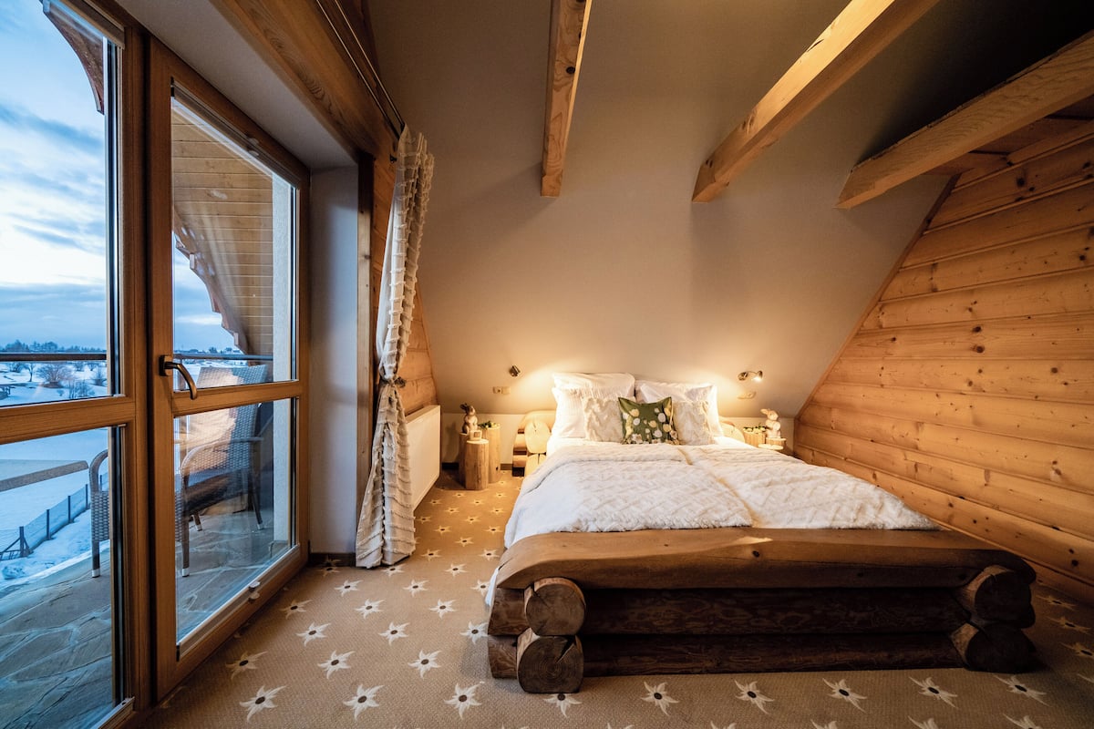 Ein gemütliches Schlafzimmer mit großem Fenster, Holzbalken, einem Bett mit weißer Bettwäsche und Blick auf eine verschneite Landschaft.