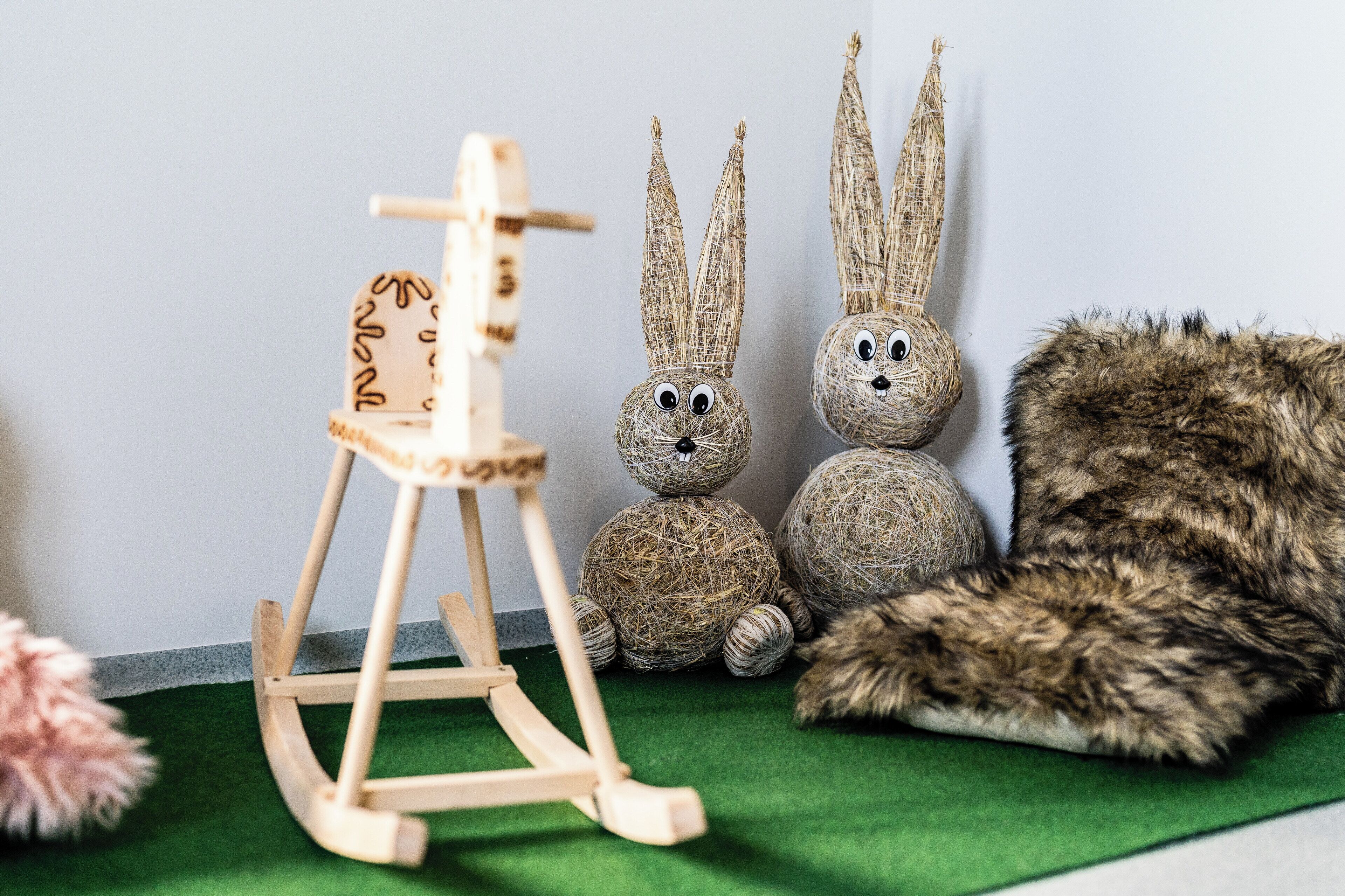 Ein hölzerner Schaukelstuhl, zwei Hasenfiguren aus Stroh und eine Stoffkatze auf einer grünen Fläche.