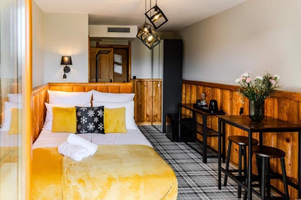 Ein Hotelzimmer mit Bett, Holz-Kopfteil, kleinem Tisch mit Hocker, Lampe und Blumen in einer Vase.