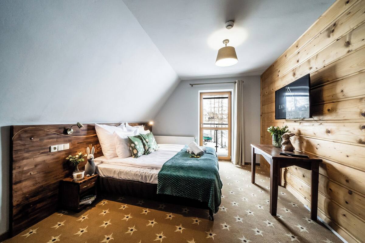 Ein Schlafzimmer mit Holz-Kopfteil, einem Bett mit weißer Bettwäsche, einem Nachttisch mit Lampe, einem kleinen Tisch mit Pflanze und einem Fenster mit Vorhängen.
