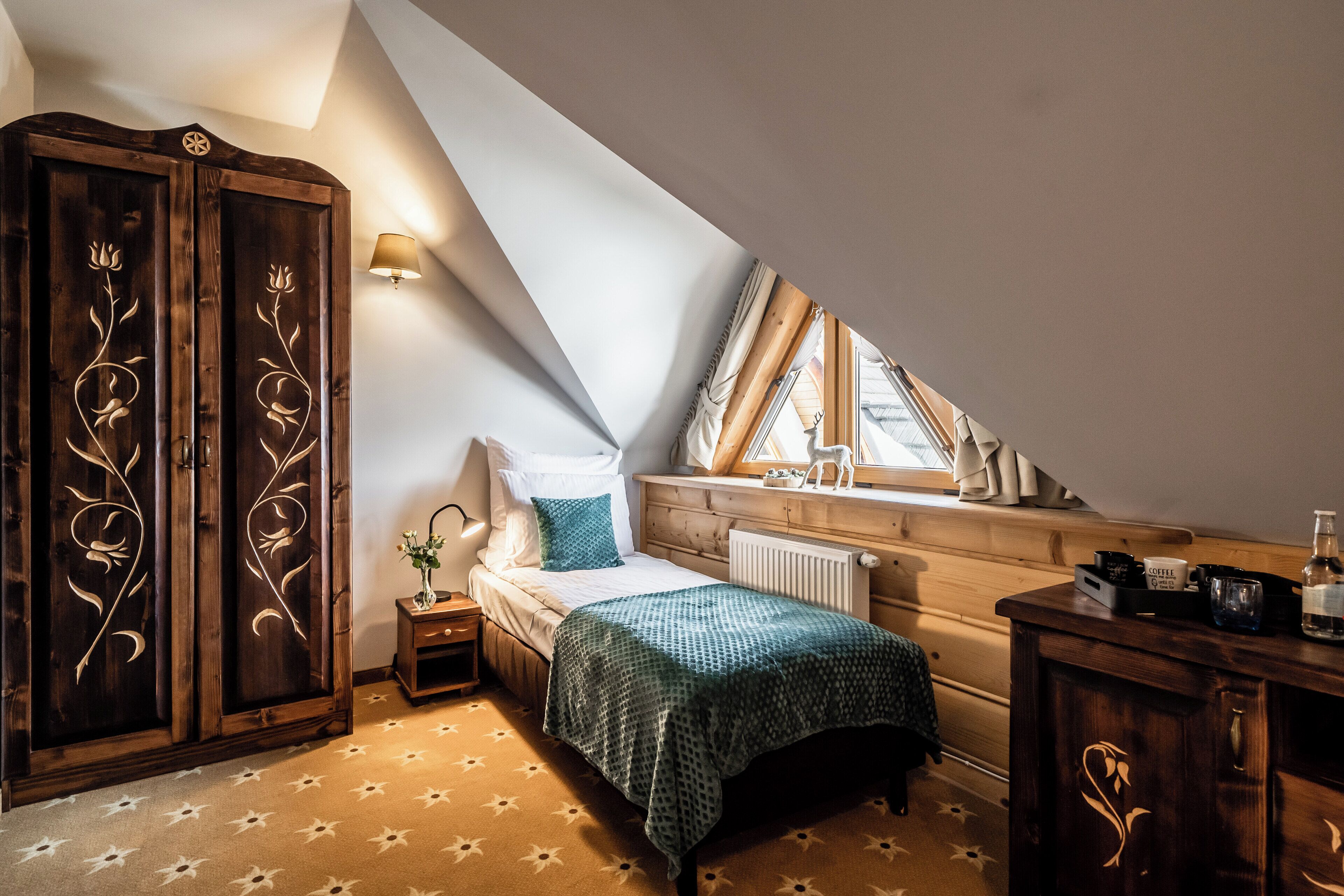 A bedroom with a sloped ceiling, a bed with a green blanket, a wooden wardrobe with floral carvings, a bedside table with a lamp, and a window with curtains.