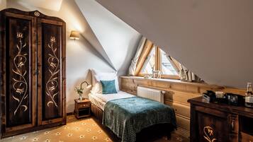 A bedroom with a sloped ceiling, a bed with a green blanket, a wooden wardrobe with floral carvings, a bedside table with a lamp, and a window with curtains.
