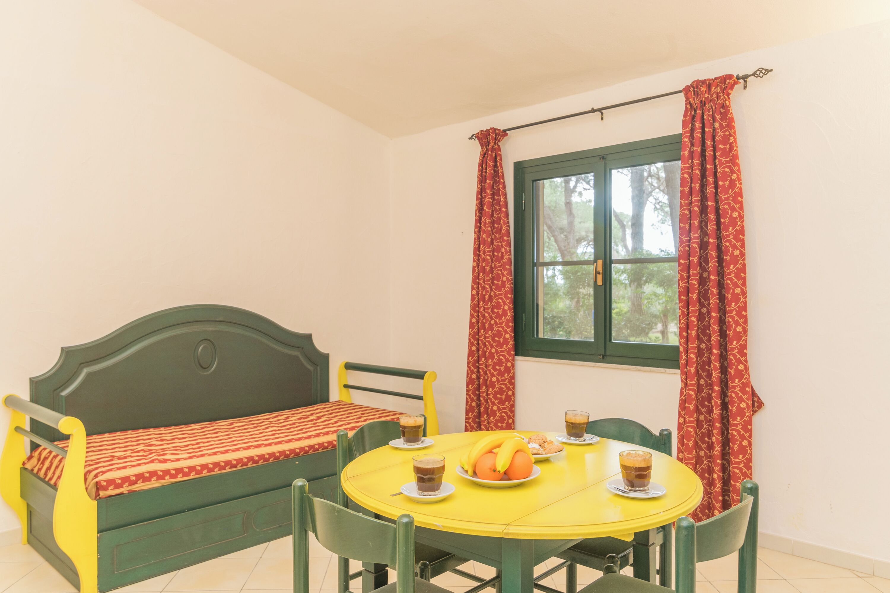 Una habitación con una cama verde, una mesa amarilla con tazas y fruta, y cortinas rojas.