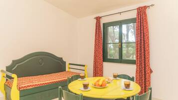 Una habitación con una cama verde, una mesa amarilla con tazas y fruta, y cortinas rojas.