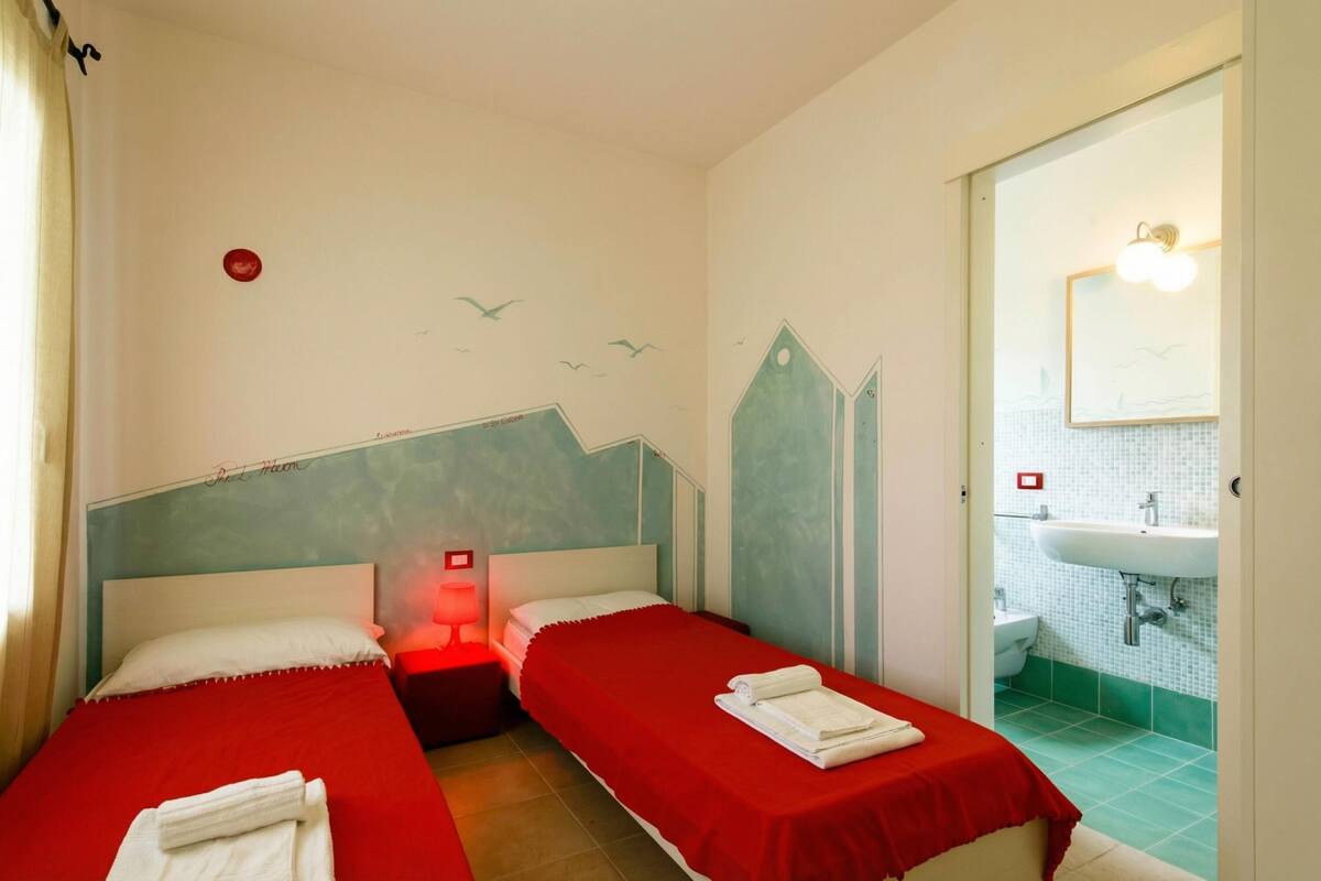 Une chambre d’hôtel avec deux lits simples, une table de chevet avec une lampe, une salle de bain avec un lavabo et un miroir, et un mur orné d’une fresque représentant des oiseaux.